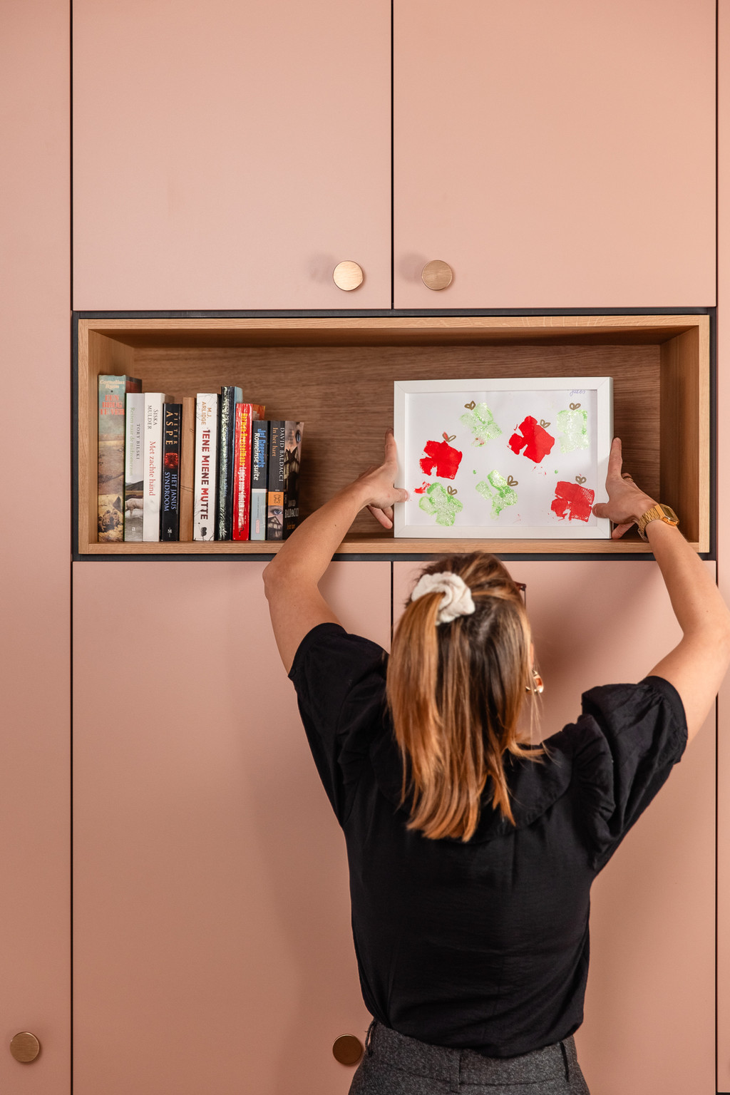 Une femme place un dessin d’enfant encadré dans la niche ouverte d’une armoire rose sur mesure. Le meuble allie fonctionnalité et touche personnelle pour un intérieur chaleureux.