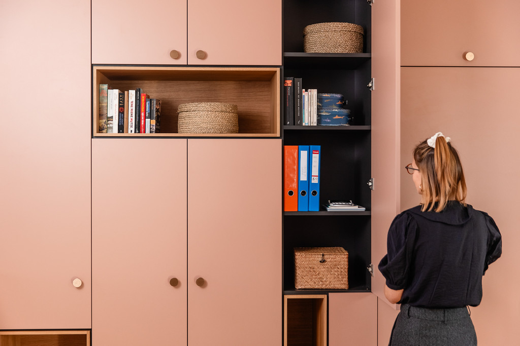 Une femme observe une armoire rose sur mesure organisée avec des livres, des classeurs et des paniers. Le meuble associe design contemporain et fonctionnalité pour un intérieur ordonné.