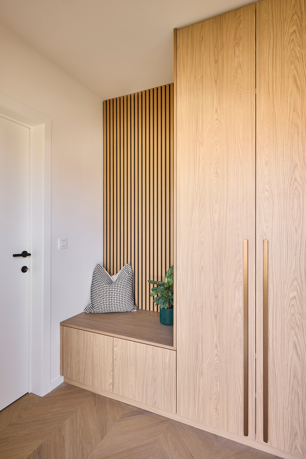 Maatwerk vestiaire in licht hout met lange geïntegreerde handgrepen, zitbank met opbergruimte en houten lattenwand, voor een warme en elegante inkomhal