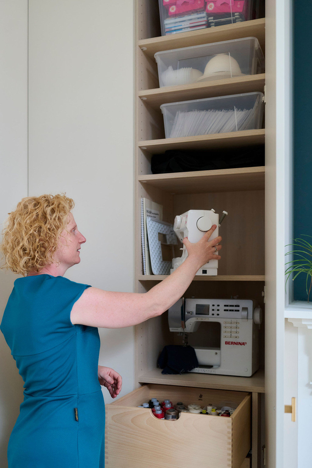 Femme qui range une machine à coudre dans un placard Camber sur mesure dédié aux loisirs, avec étagères réglables et tiroirs intégrés dans la chambre d’amis d’Els et Kevin