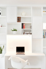 Bureau intégré dans une bibliothèque blanche avec éclairage en dessous, une chaise design, et un ordinateur portable posé sur le bureau.