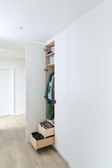 Vestiaire fermé sur mesure avec portes blanches ouvertes, étagères et tiroirs en bois avec des vêtements et accessoires.
