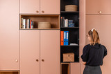 Une femme observe une armoire rose sur mesure organisée avec des livres, des classeurs et des paniers. Le meuble associe design contemporain et fonctionnalité pour un intérieur ordonné.