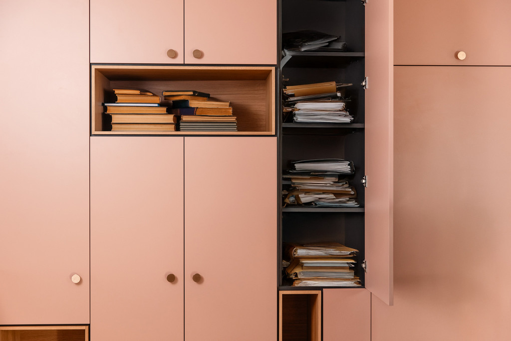 Une armoire rose sur mesure avec des dossiers et des livres empilés de manière désordonnée. Les compartiments ouverts et fermés montrent la capacité de rangement avant l’organisation finale.