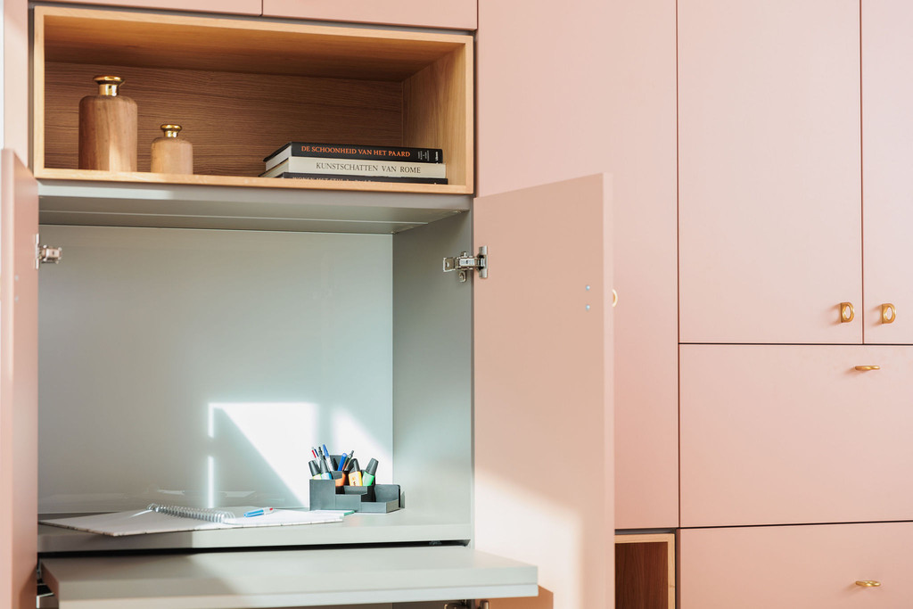 Bureau pliable sur mesure intégré dans un meuble rose, avec étagères ouvertes et accessoires de bureau comme des stylos et des carnets.