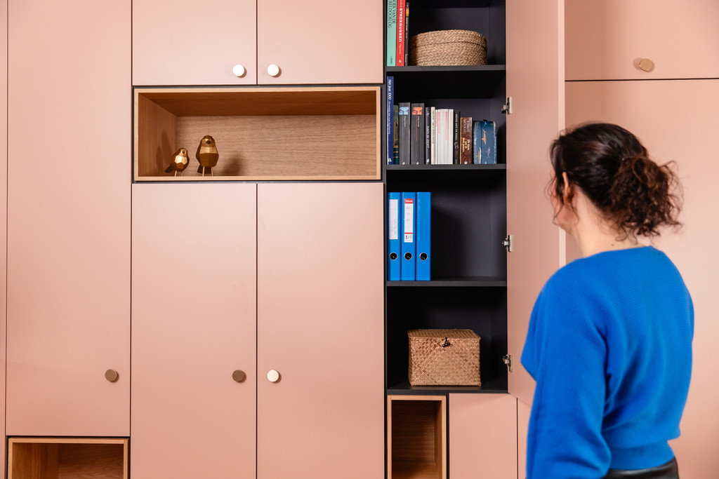 Een vrouw kijkt naar een roze maatkast met open nissen, blauwe mappen en houten accenten. De kast combineert design en functionaliteit voor een ordelijke leefruimte.