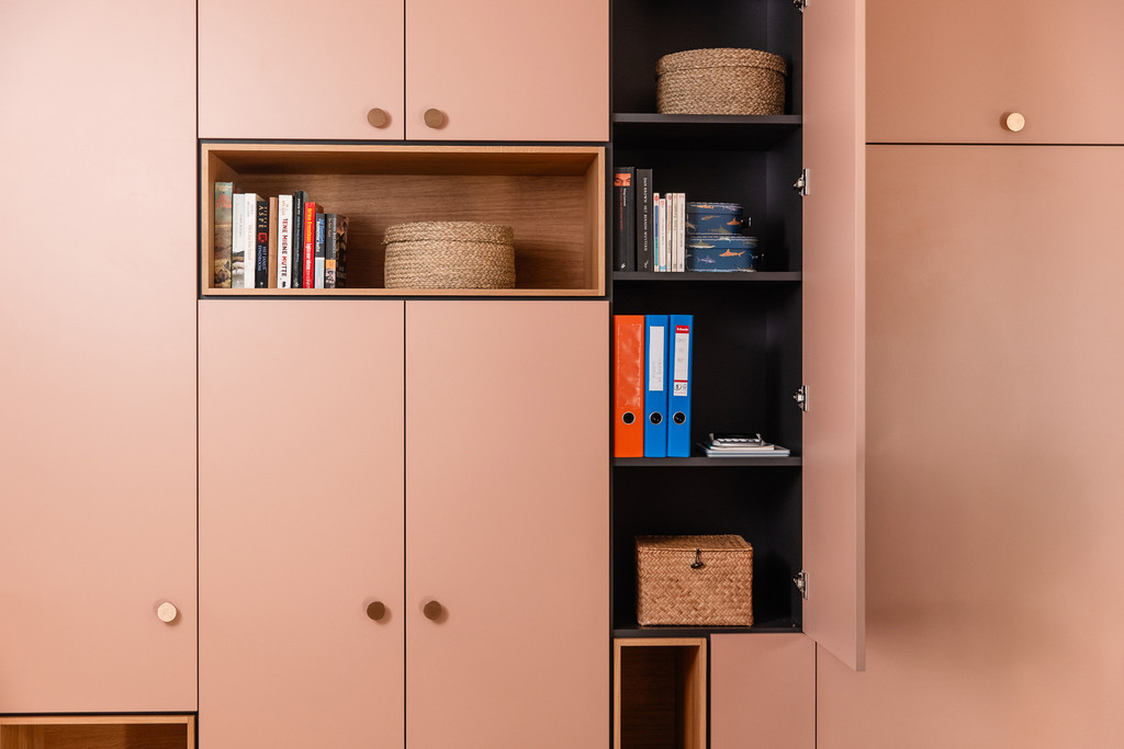 Une armoire rose sur mesure parfaitement organisée, avec des livres, des classeurs et des paniers en osier. Le meuble allie élégance et praticité pour un espace de rangement harmonieux et ordonné.