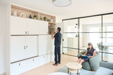 Man bergt platen op in een ingebouwde witte maatkast, vrouw leest een boek in een moderne leefruimte met glazen wanden.