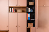 Une armoire rose sur mesure parfaitement organisée, avec des livres, des classeurs et des paniers en osier. Le meuble allie élégance et praticité pour un espace de rangement harmonieux et ordonné.