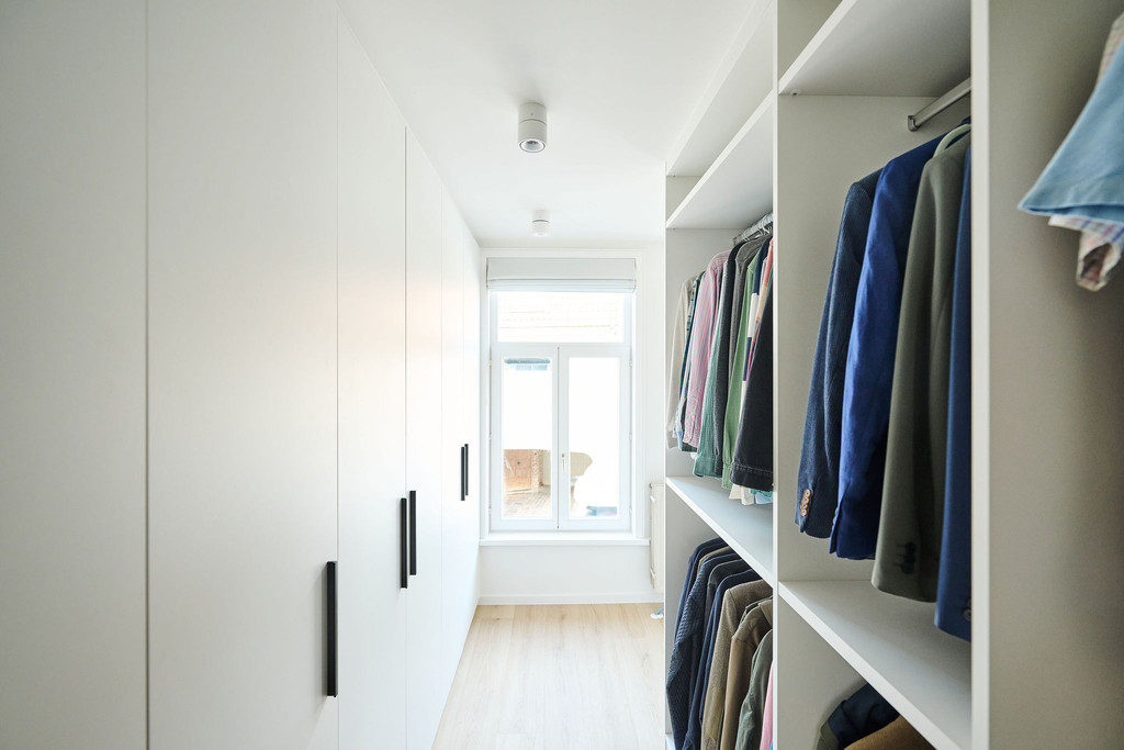 Witte dressing met gesloten maatkasten en open leg- en hangruimte langs raam in een lichte smalle ruimte