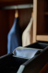 Tiroir noir ouvert avec une cravate bleue pliée dans un dressing en bois sombre, avec des costumes suspendus en arrière-plan.