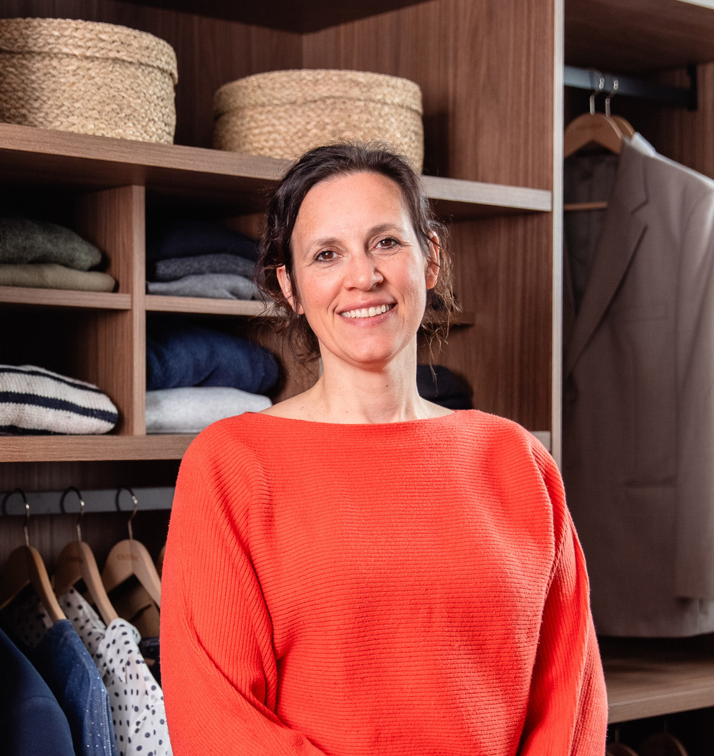 Een interieurarchitect poseert in een op maat gemaakte dressing in hout. De kast bevat open vakken met gevouwen kledij en manden, en creëert een warme en georganiseerde sfeer.