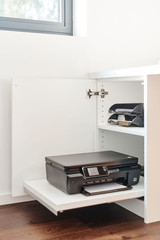 Imprimante noire placée sur une tablette coulissante intégrée dans un meuble blanc avec étagères de rangement.