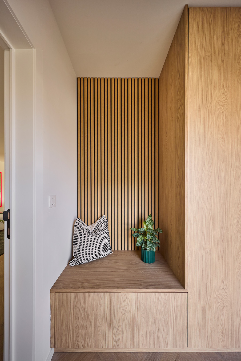 Maatwerk zithoek met houten zitbank, decoratief kussen en verticale houten lattenwand, voor een warme en uitnodigende inkomhal, gerealiseerd door Camber