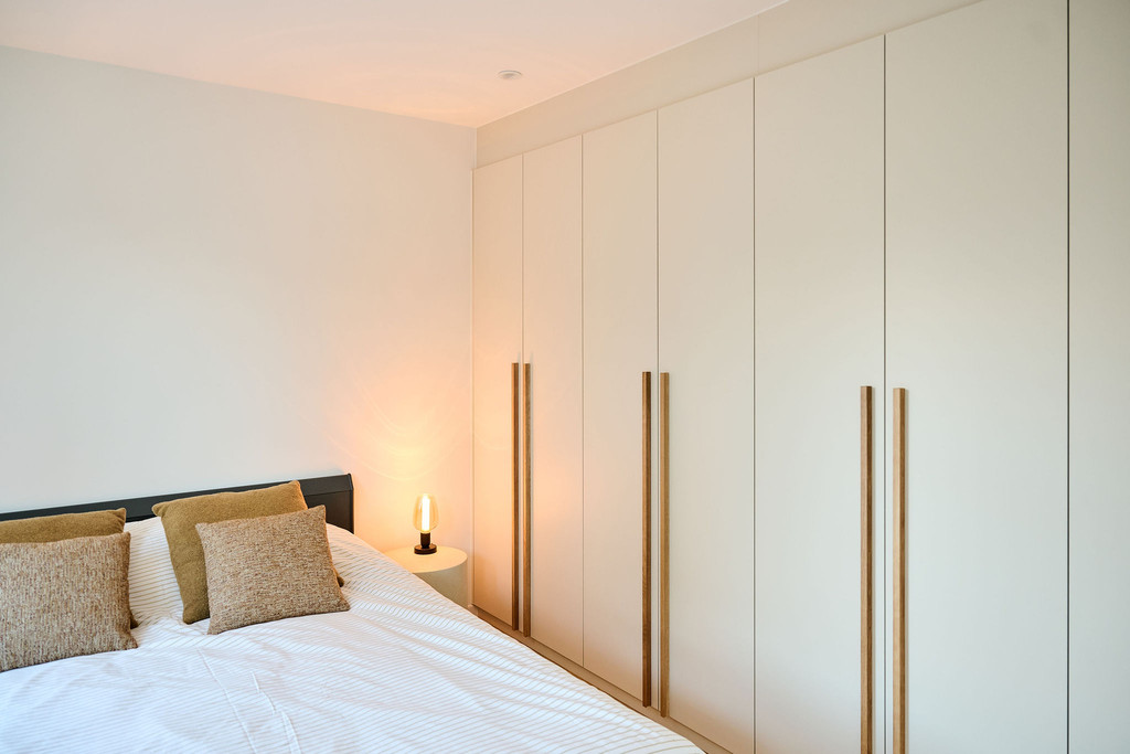 Slaapkamer met grote beige maatkast met verticale houten grepen en tweepersoonsbed met kussens