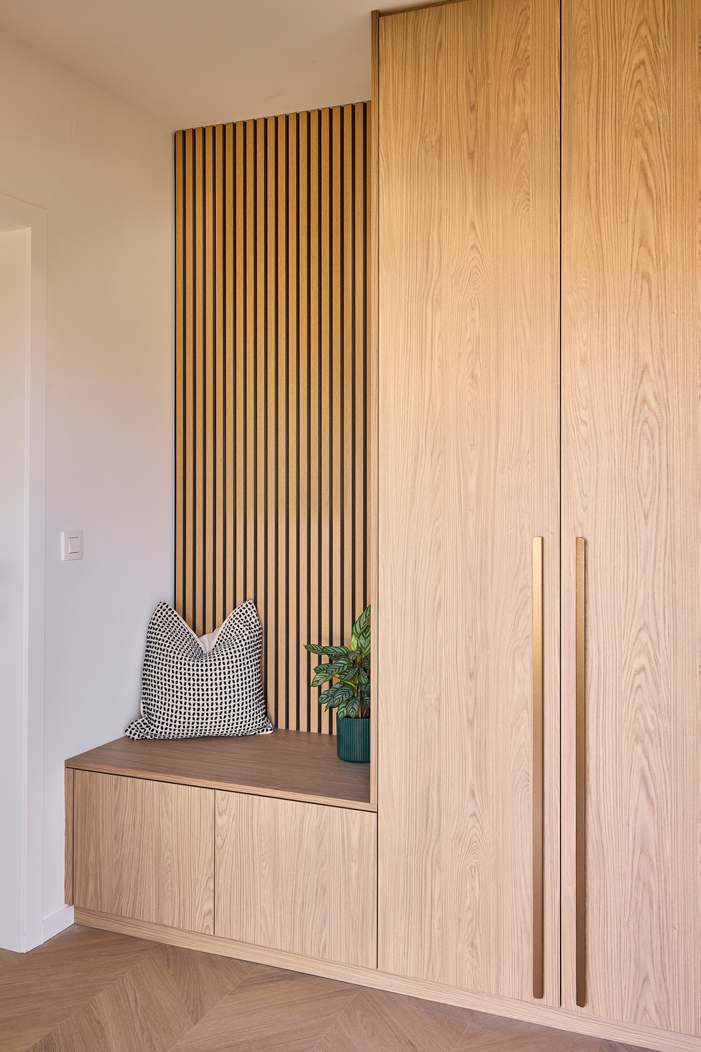 Vestiairekast op maat in licht hout met geïntegreerde zitbank, lange verticale handgrepen en houten lattenwand, voor een warme en elegante inkomhal