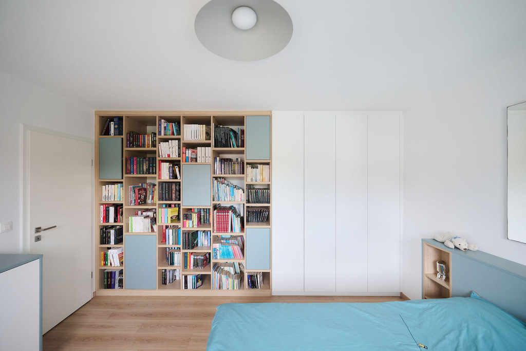 Logeerkamer met wandvullende boekenkast in hout en lichtblauw, gecombineerd met een witte kledingkast op maat en een bed met geïntegreerde opbergruimte