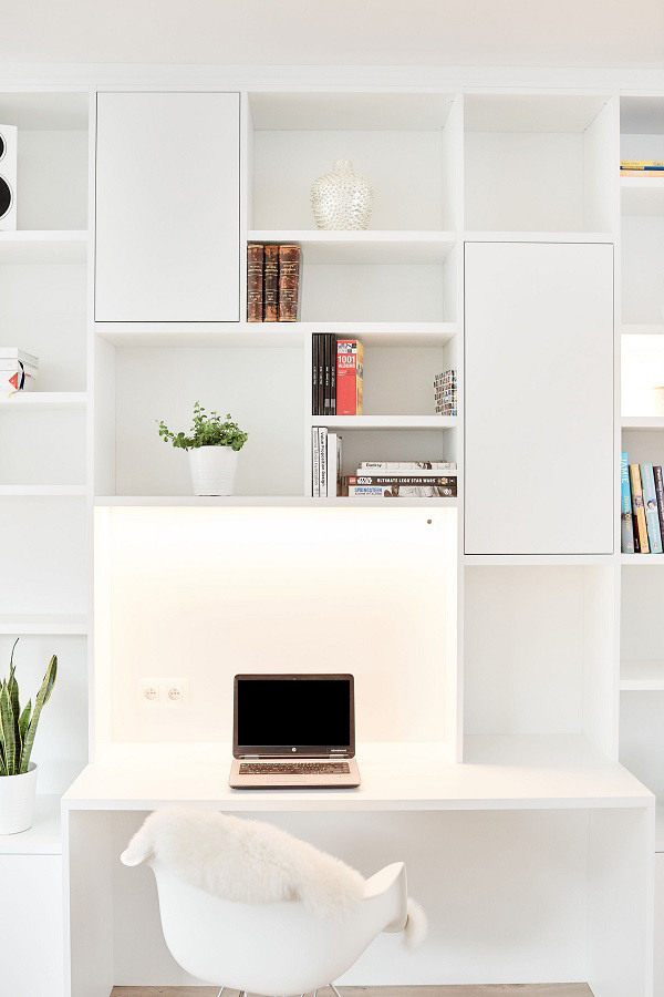 Bureau intégré dans une bibliothèque blanche avec éclairage en dessous, une chaise design, et un ordinateur portable posé sur le bureau.