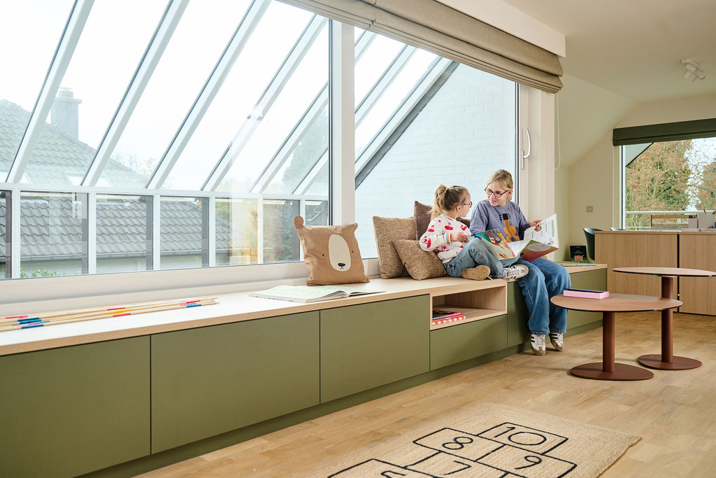 Deux enfants lisent sur une banquette verte sur mesure placée sous une grande fenêtre avec rangements intégrés