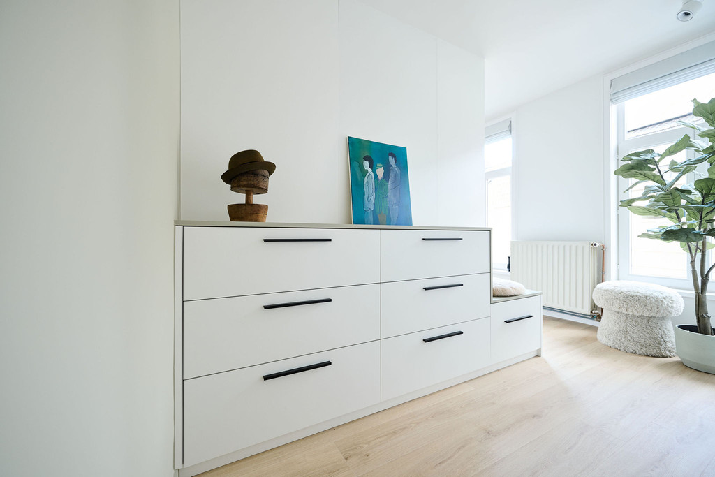 Witte ladekast op maat met zwarte grepen onder schuin dak in een lichte slaapkamer met houten vloer