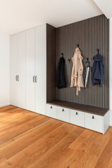 Witte ingebouwde vestiairekast met zwarte handgrepen, houten zitbank met opberglades en verticale wandbekleding met haken voor jassen en accessoires.