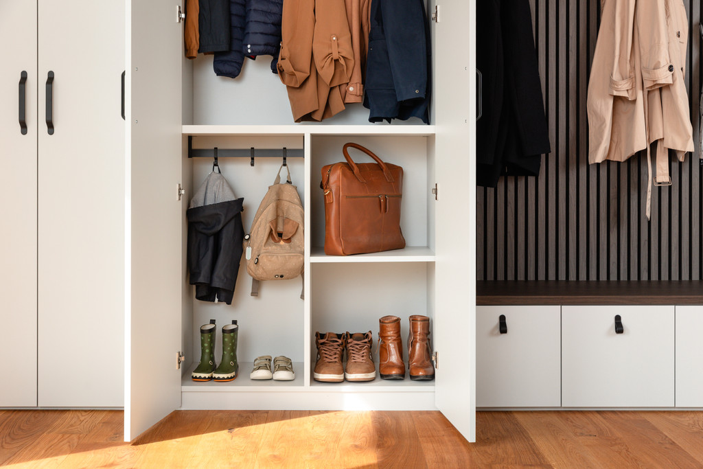 Open witte vestiairekast op maat met jassen aan een kledingroede, vakken voor rugzakken, handtas en laarzen, en een warme houten vloer.