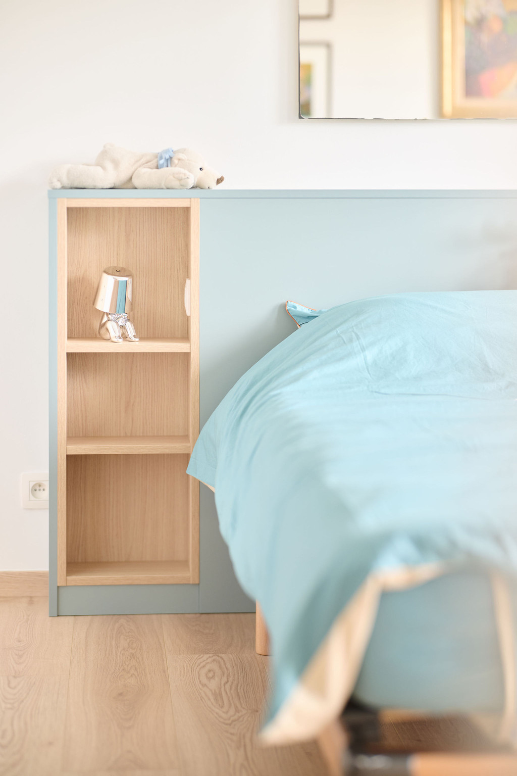 Detail van een bed op maat met lichtblauw hoofdeinde en geïntegreerd houten nachtkastje met open opbergvakken