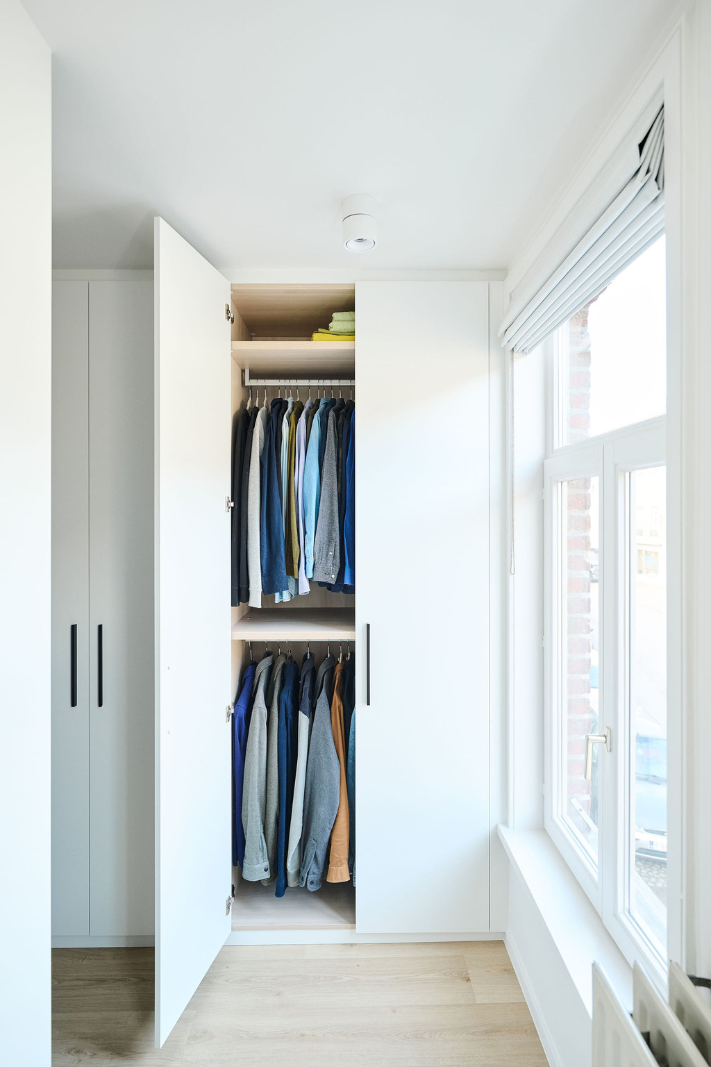 Witte ingebouwde kast met geopende deuren en dubbele hangruimte naast groot raam in een heldere dressing