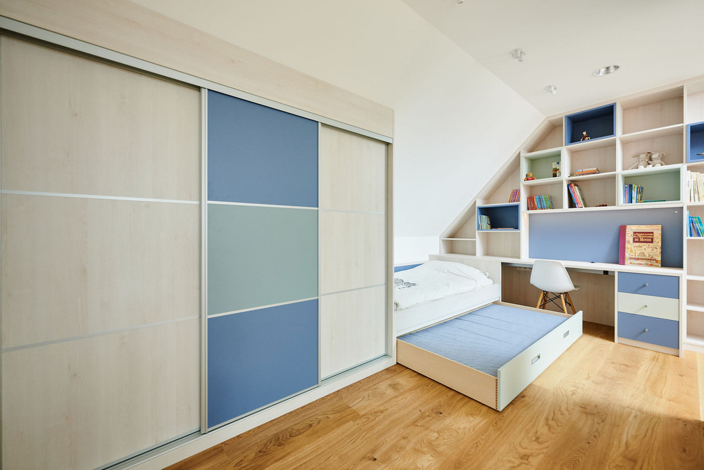 Camber Showroom Sint-Martens-Latem kinderkamer elegant stijlvol strak functionneel moderne elegant kinderkamer die goed is ingericht met slimme opbergoplossingen en een geoptimaliseerde ruimte. Aan de linkerkant staat een grote kledingkast met schuifdeuren, waarvan sommige van matglas zijn en andere gekleurd. Het eenpersoonsbed met een onderschuifbed eronder is ideaal voor logerende vrienden of voor extra opbergruimte. Aan de rechterkant past een op maat gemaakte opbergeenheid onder de schuine daklijn, met open schappen en gesloten compartimenten, die een mix van display- en opbergzones biedt. De zachte kleuren van de meubels contrasteren mooi met de lichte houten vloer, en de geïntegreerde studeertafel maakt het geheel af, waardoor er een functionele werk- of speelruimte ontstaat.